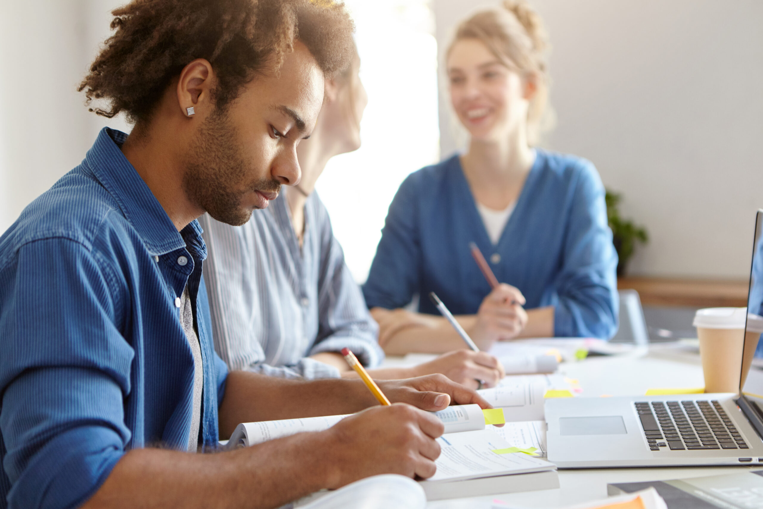 Stylish dark-skinned male in blue shirt, being busy with studying, sitting near his female groupmates, working laptop computer, writing diploma paper. Group of friendly students of different races cours-metiers-asc